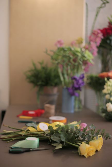 A close-up view of a workspace featuring a floral arrangement in progress, with several yellow roses, sprigs of eucalyptus, and greenery laid out on a dark grey table. In the background, there are var