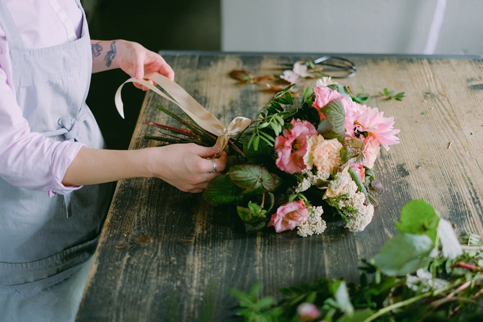 A person in a grey apron is holding a ribbon to carefully tie a floral bouquet on a rustic wooden table. The bouquet features an arrangement of fresh, pastel pink and cream flowers, including large da