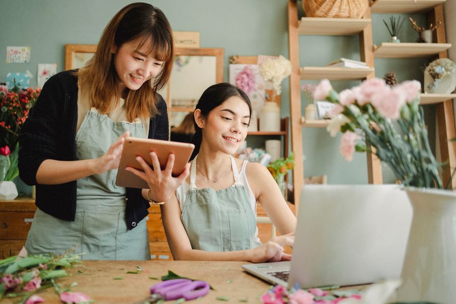 Two women in a flower shop, both wearing light green aprons, are engaged in floral arrangement activities. The woman on the left is smiling and holding a tablet, while the woman on the right is seated