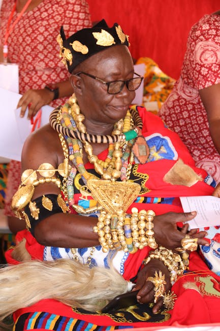 A woman dressed in traditional vibrant African attire adorned with elaborate gold jewelry including necklaces, bracelets, and a crown-like headpiece, seated indoors against bright red patterned textil